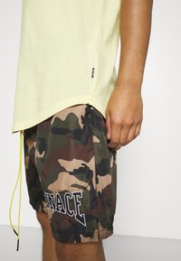 Hellgelbes Kurzarmhemd, kombiniert mit grünen und braunen Camouflage-Shorts, die ein gesticktes "Vengeance"-Detail sowie einen Kordelzug aufweisen.