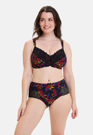 Mujer sonriente modelando un sujetador floral negro y bragas de talle alto con detalles de encaje sobre fondo blanco, mirando hacia su derecha.