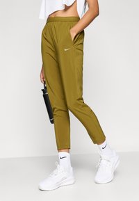 Αθλητικό παντελόνι ελιάς με ελαστική μέση και τσέπες στο πλάι, με μικρό λευκό λογότυπο Nike. Φορώντας λευκά αθλητικά παπούτσια και κάλτσες.