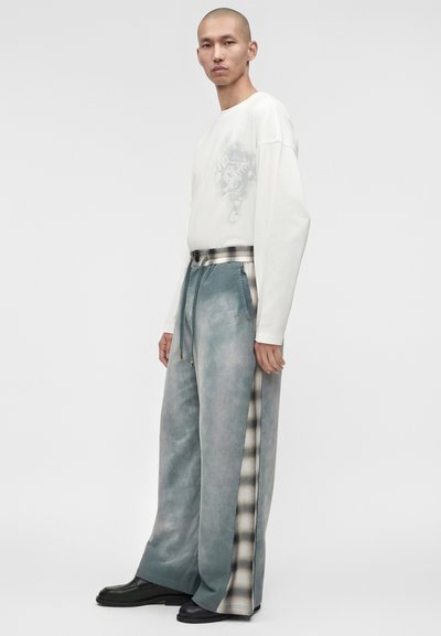 Άνδρας που στέκεται φοράει λευκό μακρυμάνικο πουκάμισο με ανοιχτό λουλουδάτο σχέδιο και φαρδιά, μπλε παντελόνια με καρό πλευρικές ρίγες και μαύρα παπούτσια.