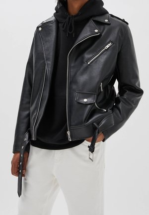 Person wearing a black leather biker jacket with silver zippers and buttons over a black hoodie and white pants, hand in pocket.