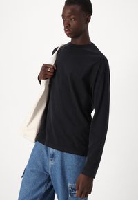 Svart langermet T-skjorte laget av bomull, med rund hals og avslappet passform. Kombinert med blå denim-bukser og holder en lyse farget totebag.
