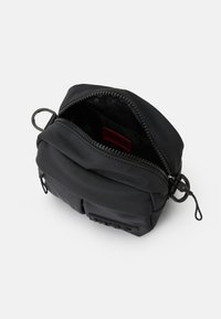 Borsa nera compatta con una zip parzialmente aperta, tasca frontale, anelli laterali e un'etichetta interna rossa con la scritta "HUGO".