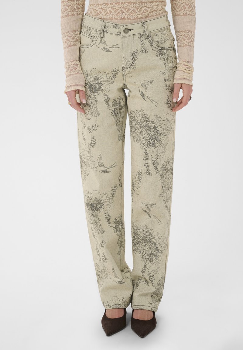 Pantalon beige droit avec des croquis noirs de fleurs et d'oiseaux, associé à un haut en dentelle à manches longues et des chaussures pointues marron foncé.