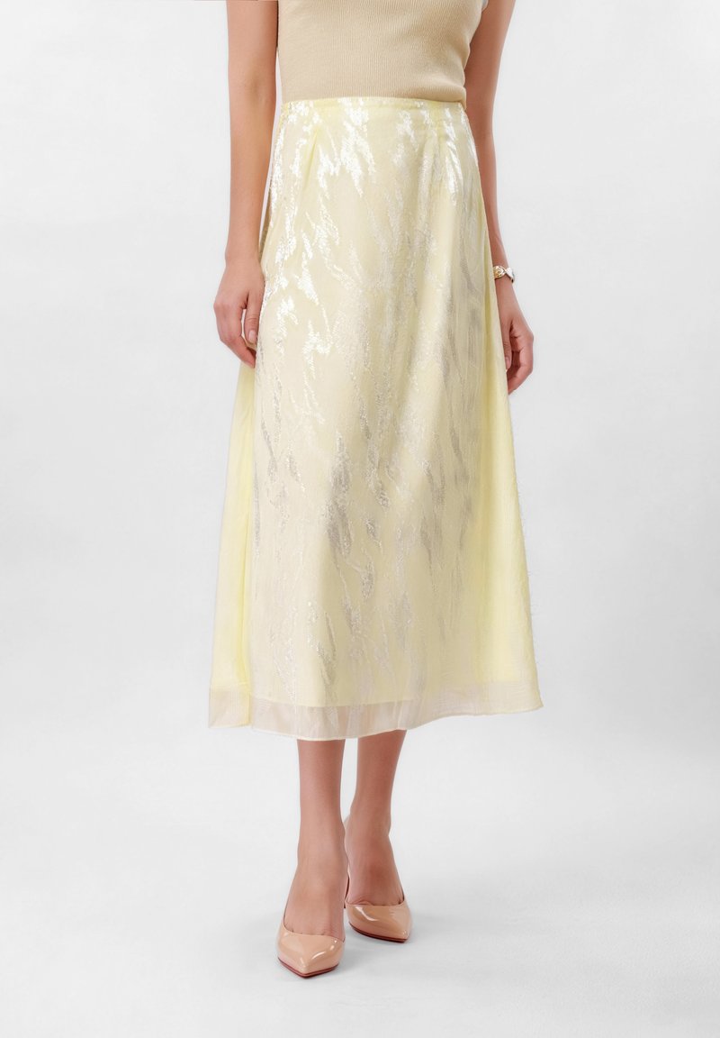 Žena v svetlo žltej sukni po lýtka so lesklým textúrovaným vzorom, béžovom bezrukávovom topu, telových špicatých podpätkoch a náramkových hodinkách.