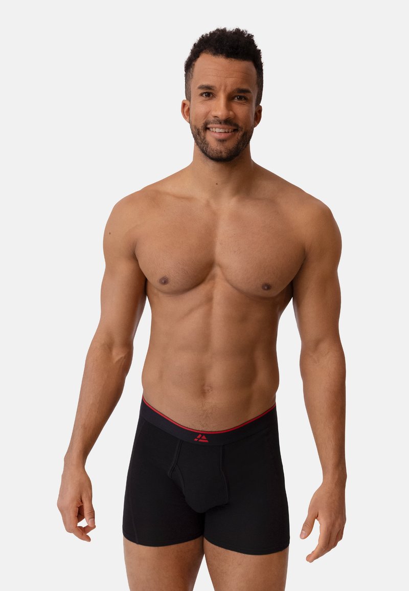 Schwarze Boxer-Briefs mit einem rot akzentuierten Bund. Weicher Stoff mit passgenauem Design und offenem Eingriff. Modell, das vor einem neutralen Hintergrund steht.