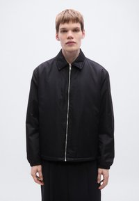 Μαύρο μπουφάν με υφή στην επιφάνεια, που διαθέτει φερμουάρ στο μπροστινό μέρος, γιακά και ελαστικές μανσέτες. Κατασκευασμένο από ελαφρύ, αδιάβροχο υλικό.