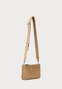 Sac bandoulière tan en matériau lisse, présentant un design à double compartiment, une sangle ajustable avec logo et des accents en matériel argenté.