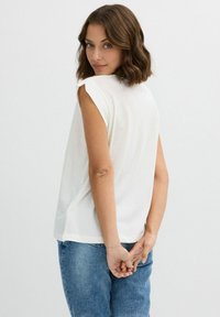 Top blanco de manga corta, holgado, con cuello redondo, combinado con jeans de corte relajado en un tono azul claro. La persona está de pie con las manos detrás de la espalda.