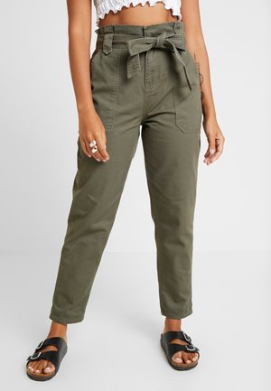 Kobieta w oliwkowozielonych spodniach z wysokim stanem i przewiązanym paskiem, białym krótkim topie, czarnych sandałach z klamrą, srebrnych pierścionkach i widocznym tatuażu na ramieniu.