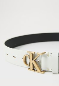 Cinturón de cuero blanco con interior negro, que presenta una gran hebilla dorada con las letras "CK" y pequeños agujeros para un ajuste ajustable.