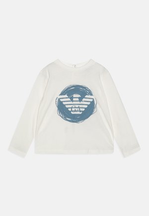 Chemise blanche à manches longues avec col rond, avec un coup de pinceau circulaire bleu et un logo d'aigle stylisé avec "GA" sur le devant.