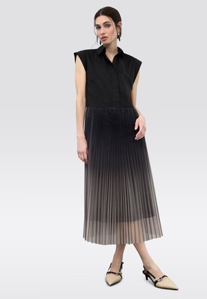 Mujer con una blusa negra sin mangas y una falda larga plisada que degrada de negro a gris, combinada con tacones beige de punta puntiaguda.