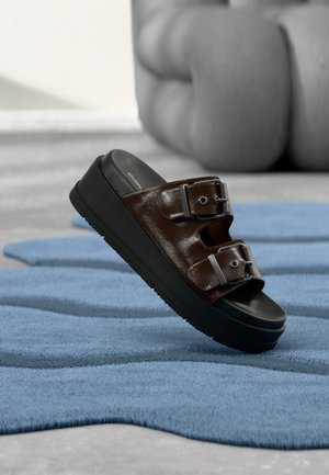Brun læder slide-sandal med to justerbare sølvspænder på en sort platformsål, hvilende på et tekstureret blåt tæppe.