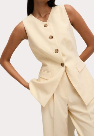 Femme portant un gilet beige sans manches avec des boutons marron et un pantalon beige à jambes larges assorti, debout les mains sur les hanches.
