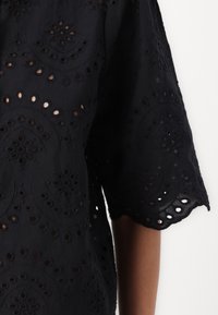 Svart brodert bluse med blomstermønster, åpne heklede detaljer og bølgete kanter på ermene. Myk kvalitet og avslappet passform.