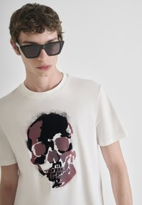 Camiseta de algodón blanca con un diseño gráfico de calavera en negro y burdeos. El modelo lleva gafas de sol negras y angulares. Textura suave.