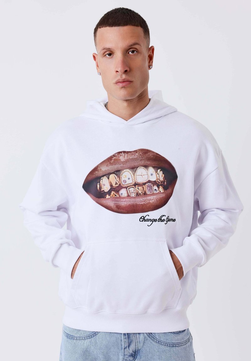 Homme aux cheveux courts portant un sweat à capuche blanc, montrant des lèvres avec des dents en or serties de bijoux et le texte « Change the Game », mains dans les poches, jean en denim clair.