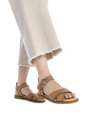 Sandalias de cuero marrón con múltiples correas, detalle de hebilla, suela plana y acentos trenzados. Combinadas con pantalones beige cortos y deshilachados.