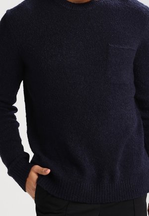 Mann trägt einen dunkelmarineblauen, strukturierten Pullover mit Brusttasche, die Hand ruht in der Tasche einer schwarzen Hose.