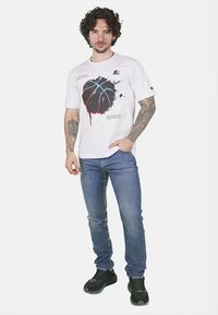 Maglietta bianca in cotone con una grafica di basket nera e rossa, linee blu e testo. Abbinata a jeans blu e scarpe da ginnastica nere.