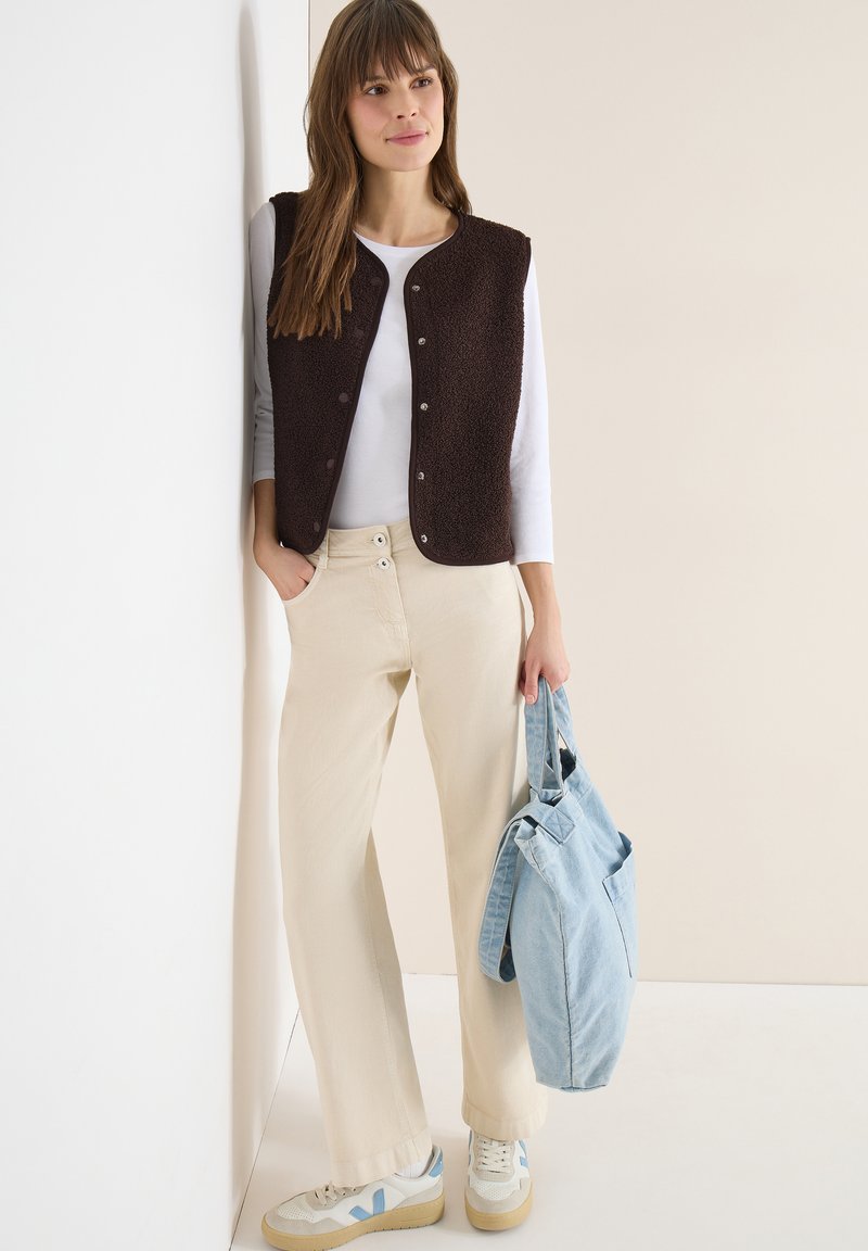 Femme appuyée contre un mur, portant une chemise blanche à manches longues, un gilet marron foncé, un pantalon beige, des baskets, et tenant un sac fourre-tout bleu clair.