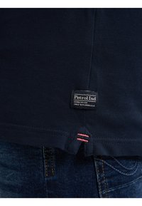 Námořnická modrá polo tričko s texturovanou tkaninou, obsahující značkovou etiketu ("Petrol Ind") a malé červené akcentní šití na lemu.