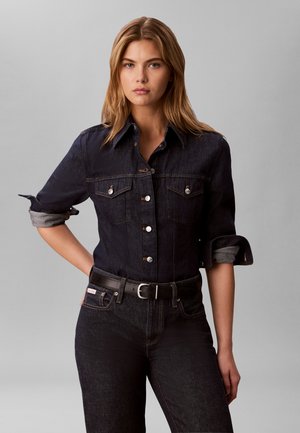 Giovane donna con capelli biondi che indossa una giacca di jeans scura e jeans, in piedi con una mano sui fianchi su uno sfondo grigio uniforme.