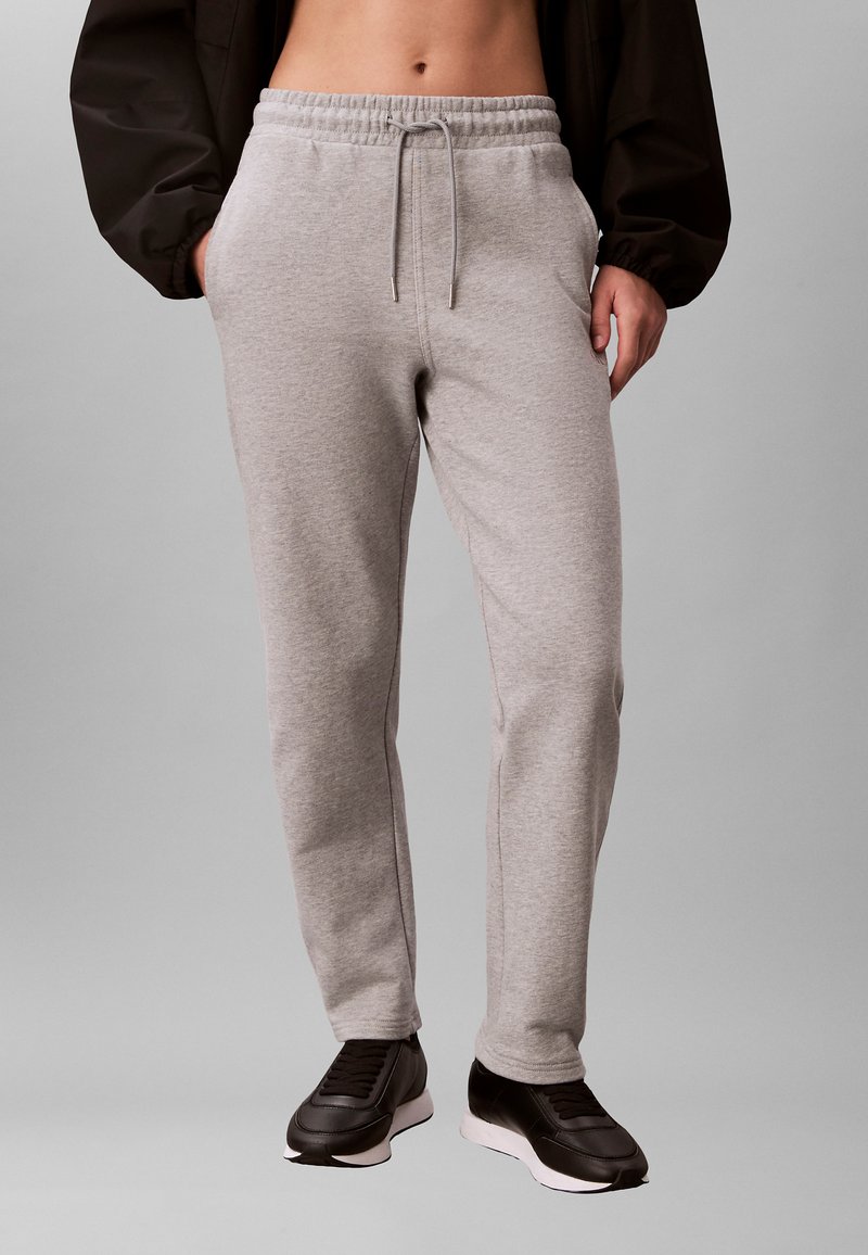 Personne portant un pantalon de survêtement gris clair avec des poches à cordon, une veste noire et des baskets noires sur un fond neutre.