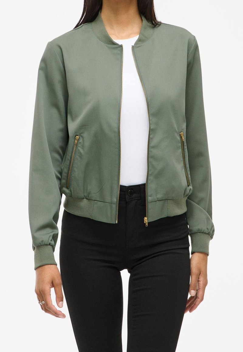 Femme portant une veste bomber vert olive avec des poches zippées dorées sur une chemise blanche et un pantalon noir, debout devant un fond blanc.
