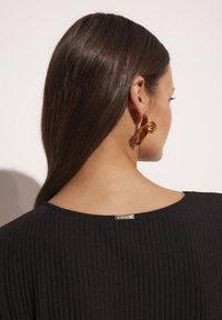 Czarny prążkowany top z okrągłym dekoltem, z metalicznym akcentem z tyłu. Złote kolczyki z wyrazistym wzorem.