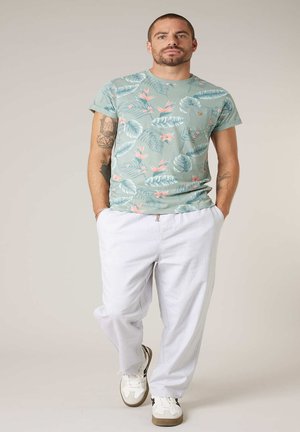 Homme marchant les mains dans les poches, portant un t-shirt vert à imprimé floral, un pantalon blanc, des baskets blanches, avec des tatouages visibles sur les bras.