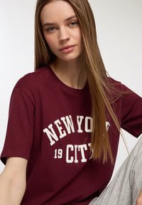 T-shirt di cotone bordeaux con maniche corte, caratterizzato da una grafica bianca "NEW YORK 19 CITY" in un carattere audace. Texture morbida, design casual.