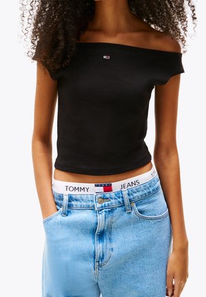 Nainen, jolla on päällään musta olkapäät paljastava lyhyt toppi ja vaaleansiniset korkeavyötäröiset farkut, joiden vyötäröllä näkyy Tommy Jeans -brändätty vyötärönauha.