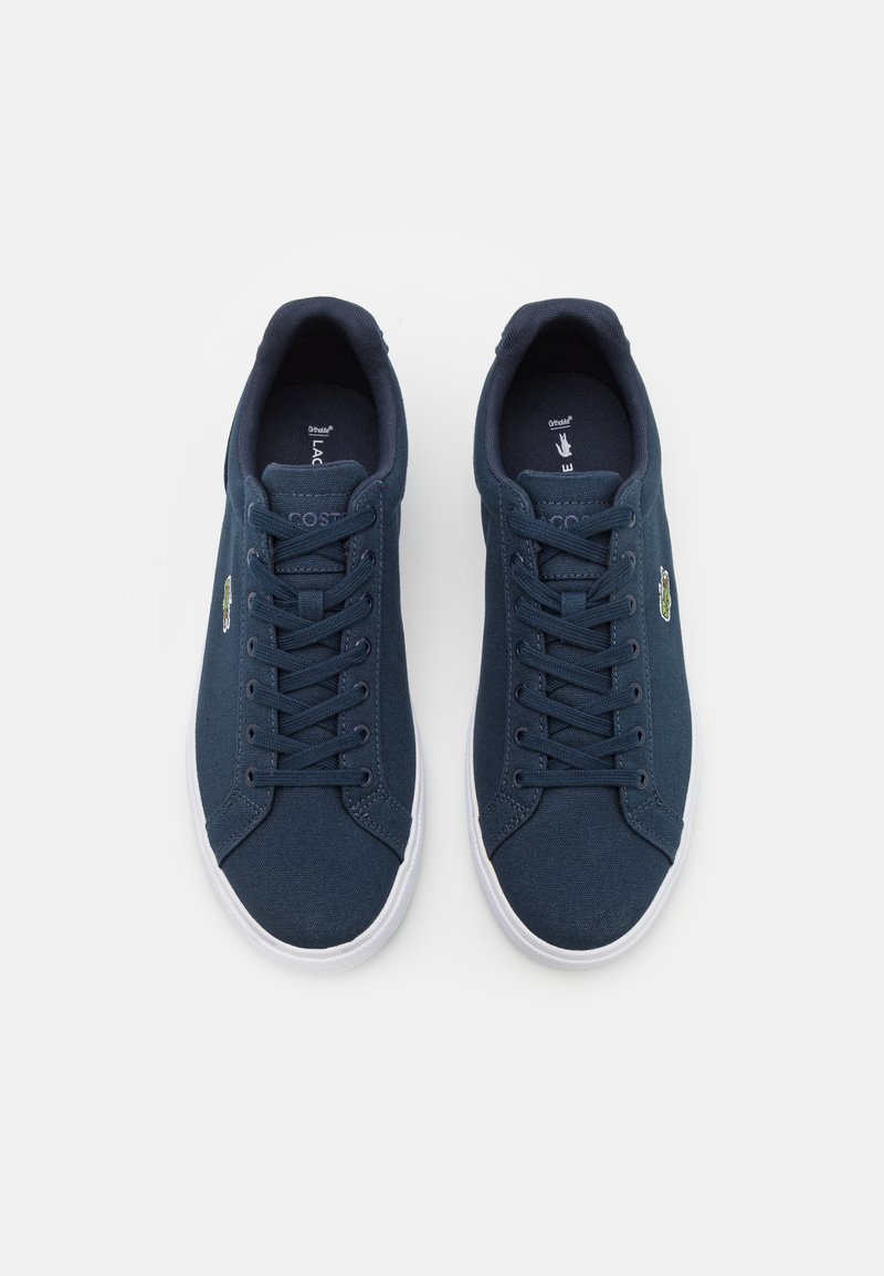 Sneakers in tela blu navy con suole in gomma bianca. Le caratteristiche includono punta tonda, design con lacci e logo del coccodrillo Lacoste sul lato.