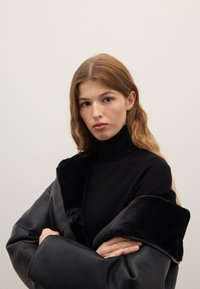 Schwarze Lederjacke mit plüschigem schwarzem Kragen, getragen über einem schwarzen Rollkragenpullover. Lange braune Haare und neutraler Hintergrund vervollständigen den Look.