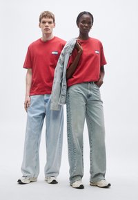 T-shirts vermelhas de algodão com pequenos patches de logótipos, combinadas com jeans de perna larga azul claro. Um modelo segura uma jaqueta de ganga e ambos usam ténis.