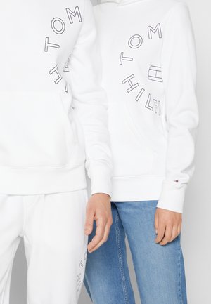 Witte hoodies met zwarte tekst die "TOMMY HILFIGER" in een cirkelontwerp lezen. De stof is glad; heeft een voork pocket en een trekkoord capuchon.