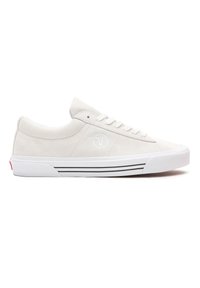 Suede lave sneakers i lys beige med hvite snørebånd, en logo på siden og sorte striper på sålene. Flat silhuett og avrundet tådesign.