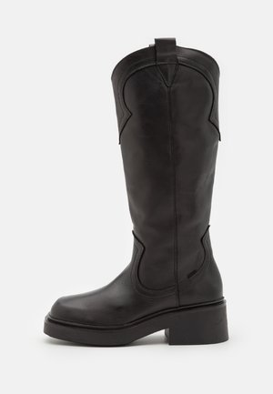 Botte haute en cuir noir avec bout arrondi, coutures décoratives, languette en haut et talon bloc épais sur fond blanc.