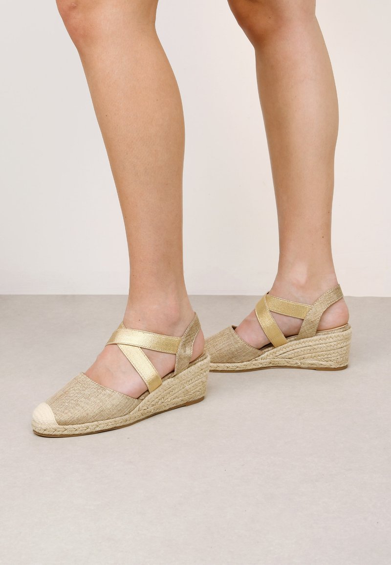Beige espadrille wedge sandals with crisscross golden straps, a textured fabric upper, and a woven jute sole. Closed cap toe detail.