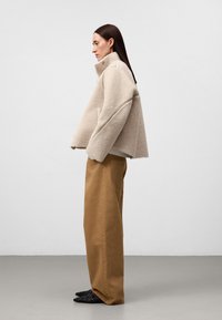 Beżowa, futrzasta kurtka o wysokim kołnierzu, w parze z luźnymi spodniami w kolorze taupe. Włosy długie i proste, stojąc bokiem na szarym podłożu.