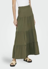Falda maxi verde oliva en capas, hecha de tela suave. Presenta una cinturilla elástica y un diseño fluido, combinada con sandalias negras.