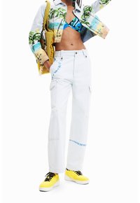 Person som bär en tropisk mönstrad kort jacka, blå mönstrad bikiniöverdel, vita cargo-byxor med blå detaljer, gula sneakers och bär en gul vävd väska.