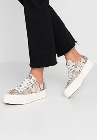 Persona che indossa pantaloni neri a taglio corto e sneakers bianche con suola alta con stampa pitone, in piedi su un pavimento grigio chiaro.