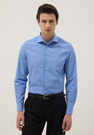 Uomo che indossa una camicia celeste con bottoni infilata in pantaloni neri con cintura nera, in piedi davanti a uno sfondo uniforme.