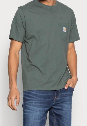 Mand iført en grøn kortærmet pocket t-shirt med Carhartt-logo og blå jeans, hånden i lommen, foran en ensfarvet baggrund.