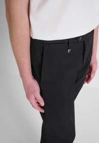 Crne hlače s krojem koji odgovara tijelu, nabranjem sprijeda i zatvaranjem na gumb i patentni zatvarač. Glatka tekstura tkanine s minimalnim detaljima.