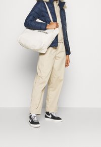 Modrá péřová bunda s kapucí lemovanou kožešinou, béžové kalhoty a černé tenisky. Nese velkou plátěnou tašku s potiskem.