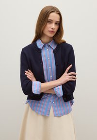 Cardigan lavorato a maglia blu navy sopra una camicia a bottoni a righe blu con righe rosa e bianche. Il cardigan ha un colletto rotondo e bottoni.